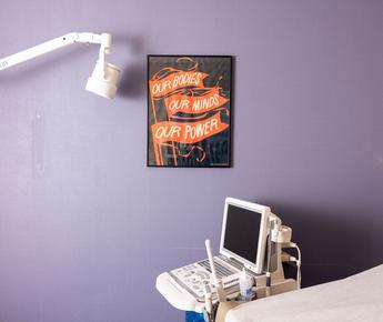 An ultrasound machine in a clinic room with a lavender wall. A sign on the wall reads "Our bodies our minds our power"