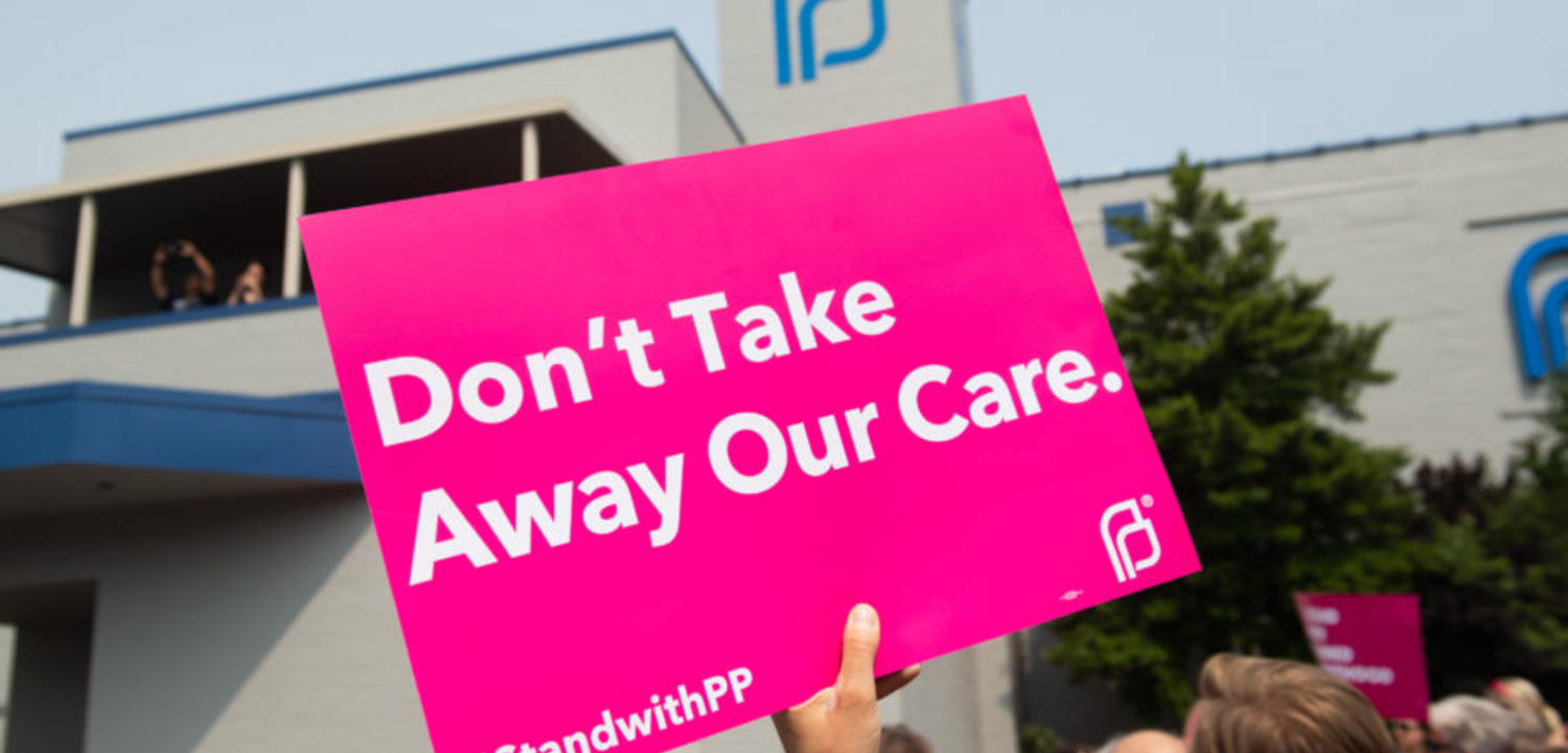 A protest at a Planned Parenthood with a pink sign reading "Don't take away our care".