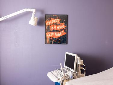 An ultrasound machine in a clinic room with a lavender wall. A sign on the wall reads "Our bodies our minds our power"