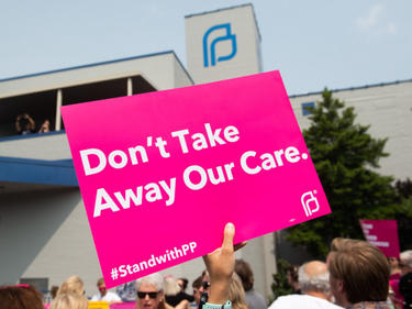 A protest at a Planned Parenthood with a pink sign reading "Don't take away our care".