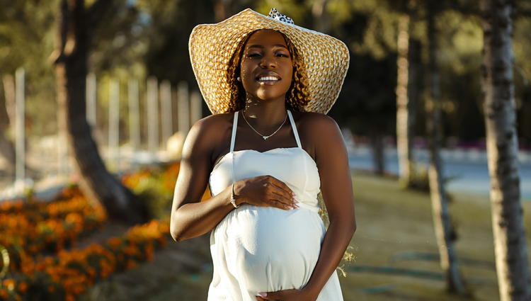 Black woman in white spaghetti strap dress wearing brown sun hat holding pregnant belly. Credit: Ben Iwara